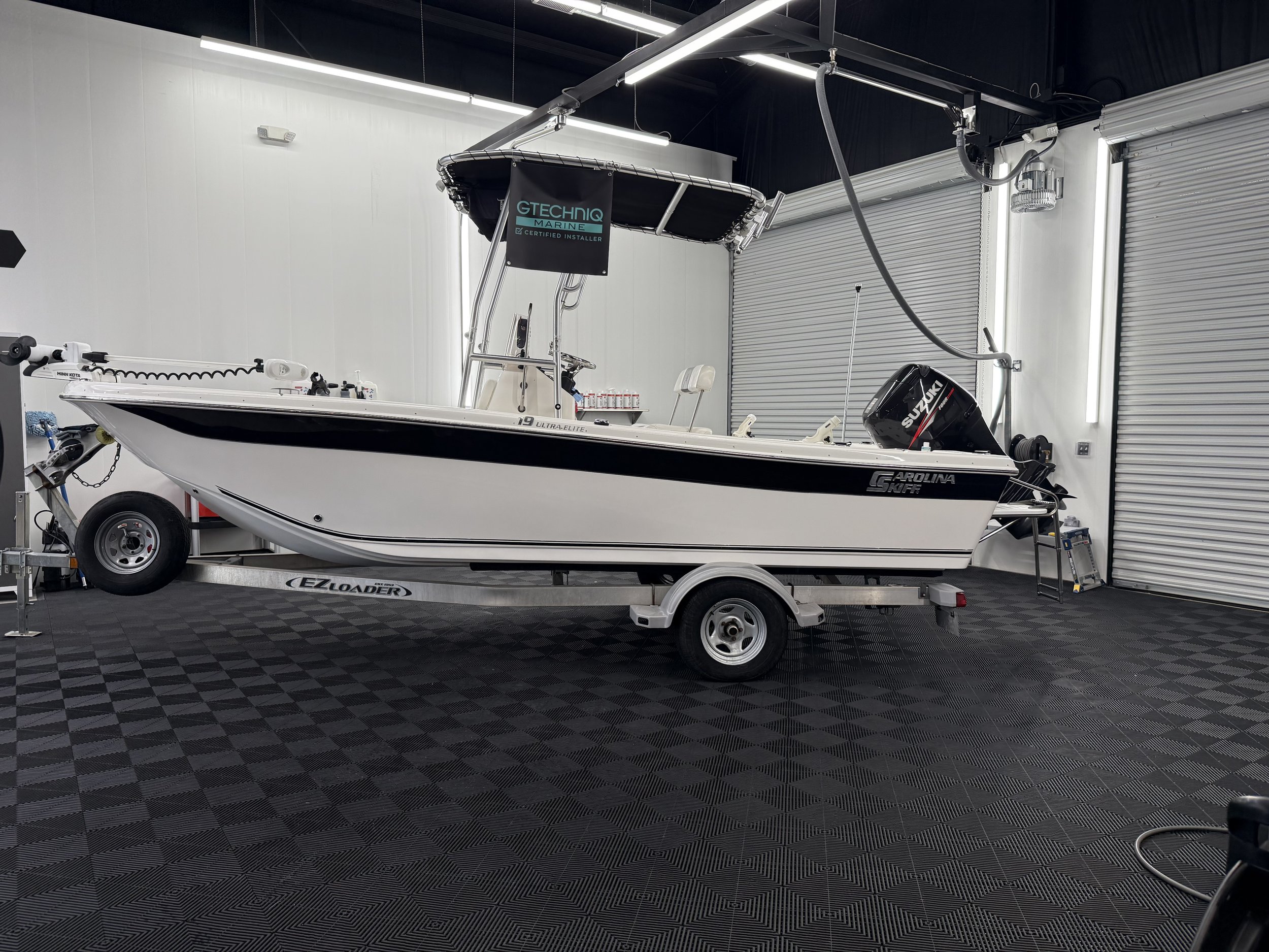 A Carolina Skiff boat on a trailer inside a workshop with black checkered flooring, white walls, and rolling metal doors.
