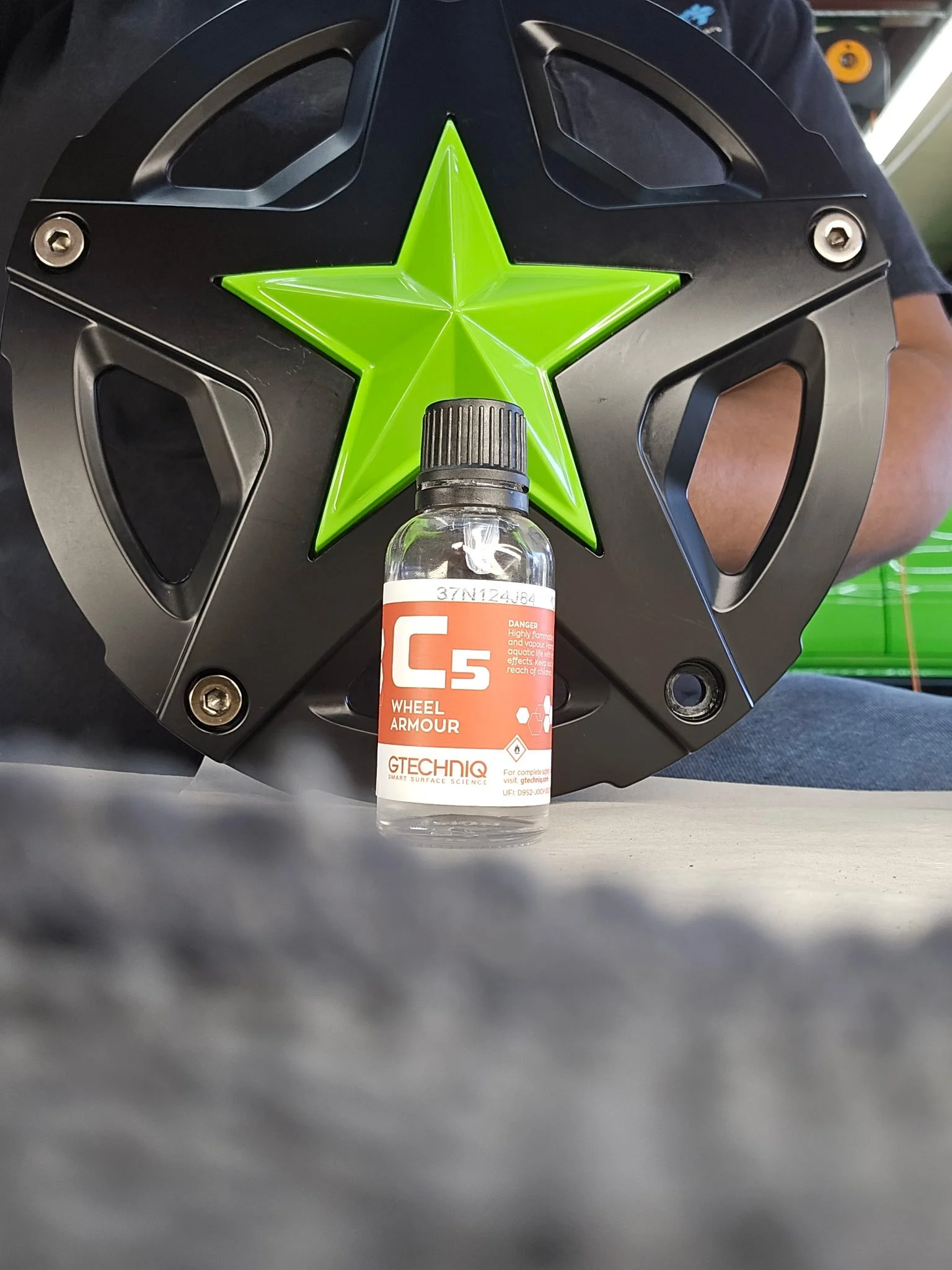 A motorcycle helmet with a green star logo and screws on the sides, placed on a table. In front of the helmet is a bottle of Gtechniq C5 Wheel Armour. A person with a dark shirt is partially visible in the background.