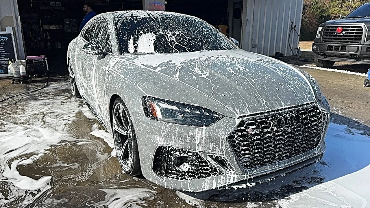 A white Audi sedan covered in soap suds during a car wash, with foam dripping down its surface, outside in front of a garage. ceramic g wash