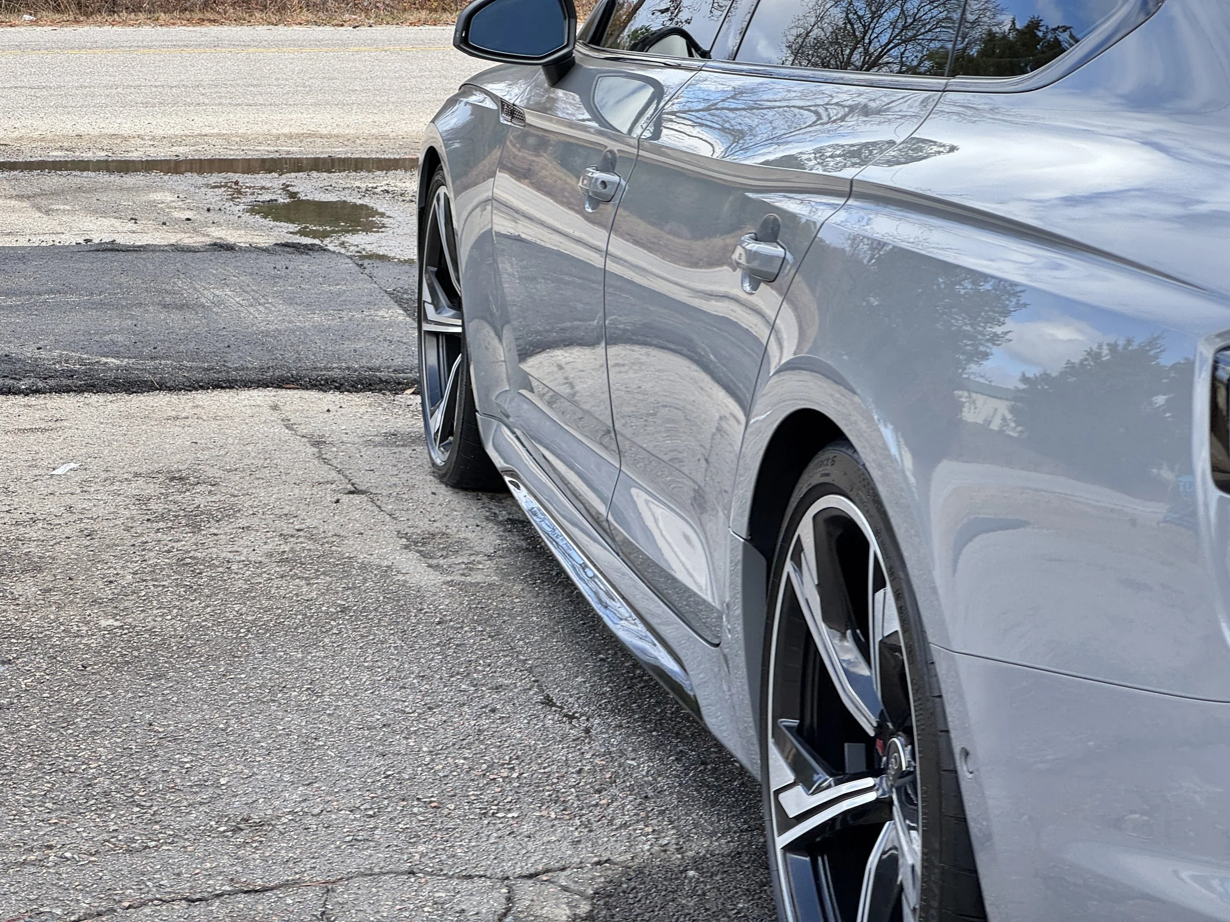 Close-up of the side of a silver luxury car parked on a cracked asphalt surface, showing detailed reflections on the body and alloy wheels with low-profile tires.
