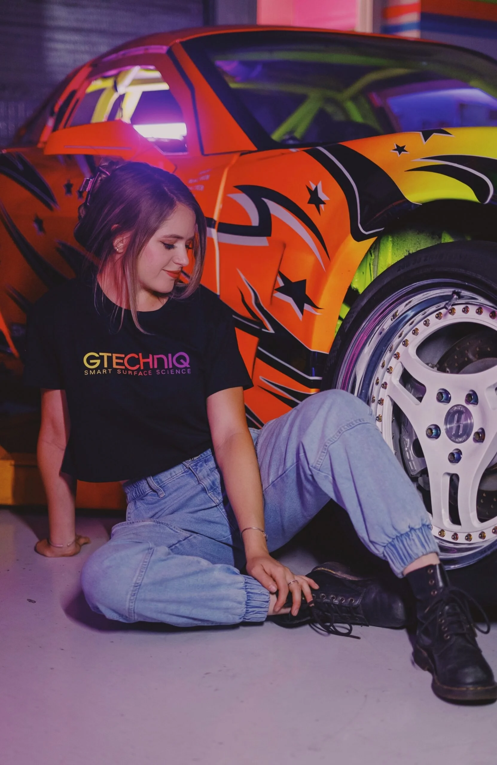 A young woman sitting on the ground in front of a colorful customized sports car, wearing a black T-shirt with 'GTECHNIQ' printed on it and light blue jeans.