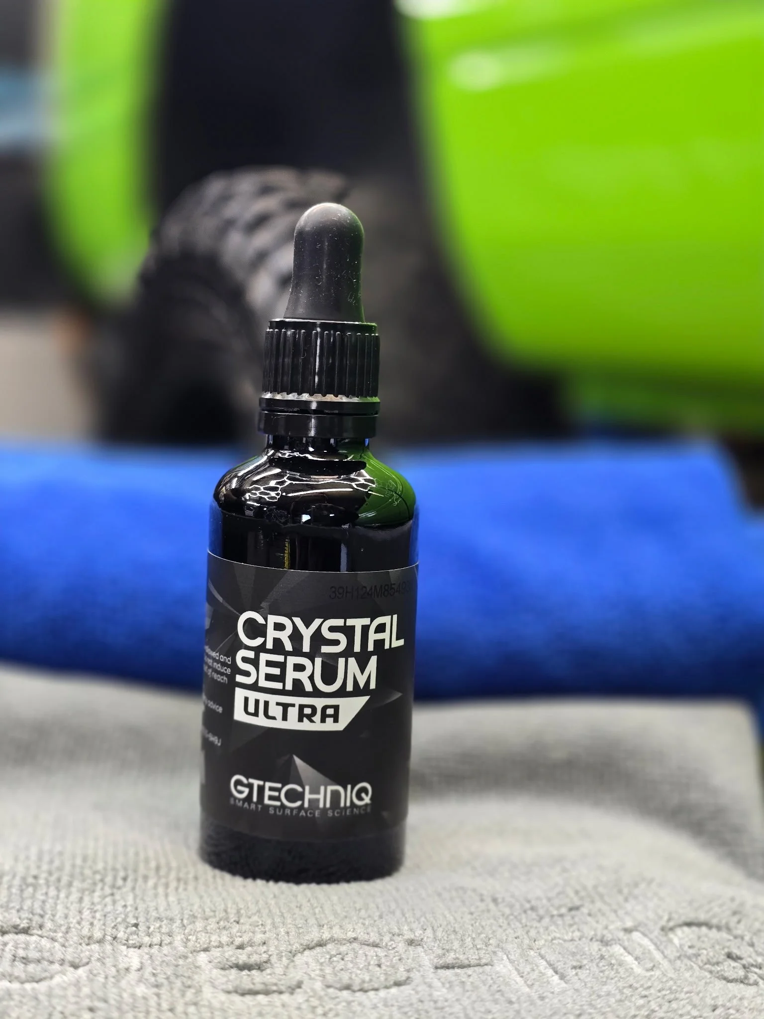 A small black bottle with a dropper cap labeled 'Crystal Serum Ultra' by GTECHNIQ, placed on a textured surface with a blurred background featuring a person wearing black shoes and green clothing.