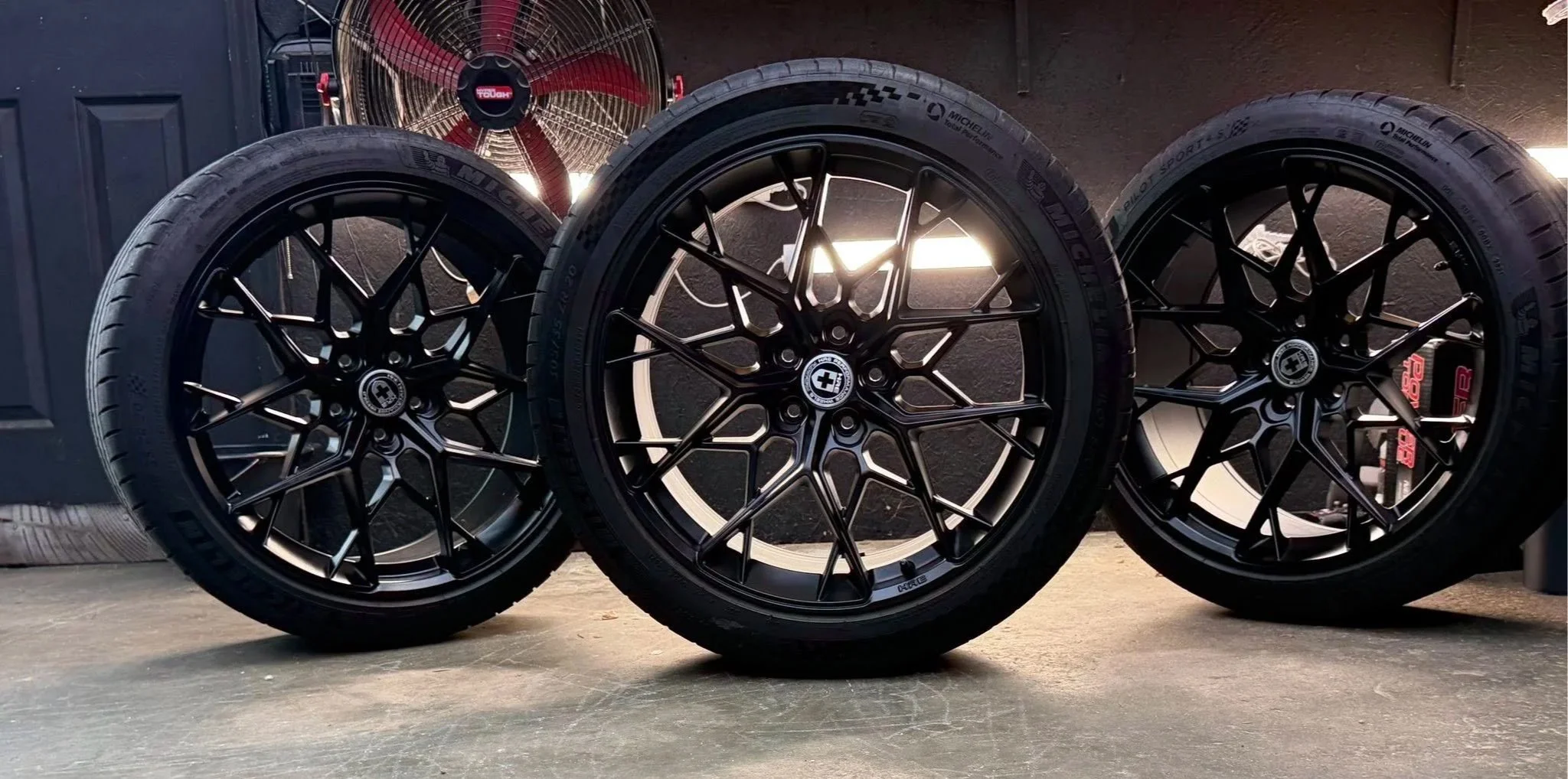 Three black alloy car wheels with tires, arranged in a row on the floor, with a fan and wall in the background.
