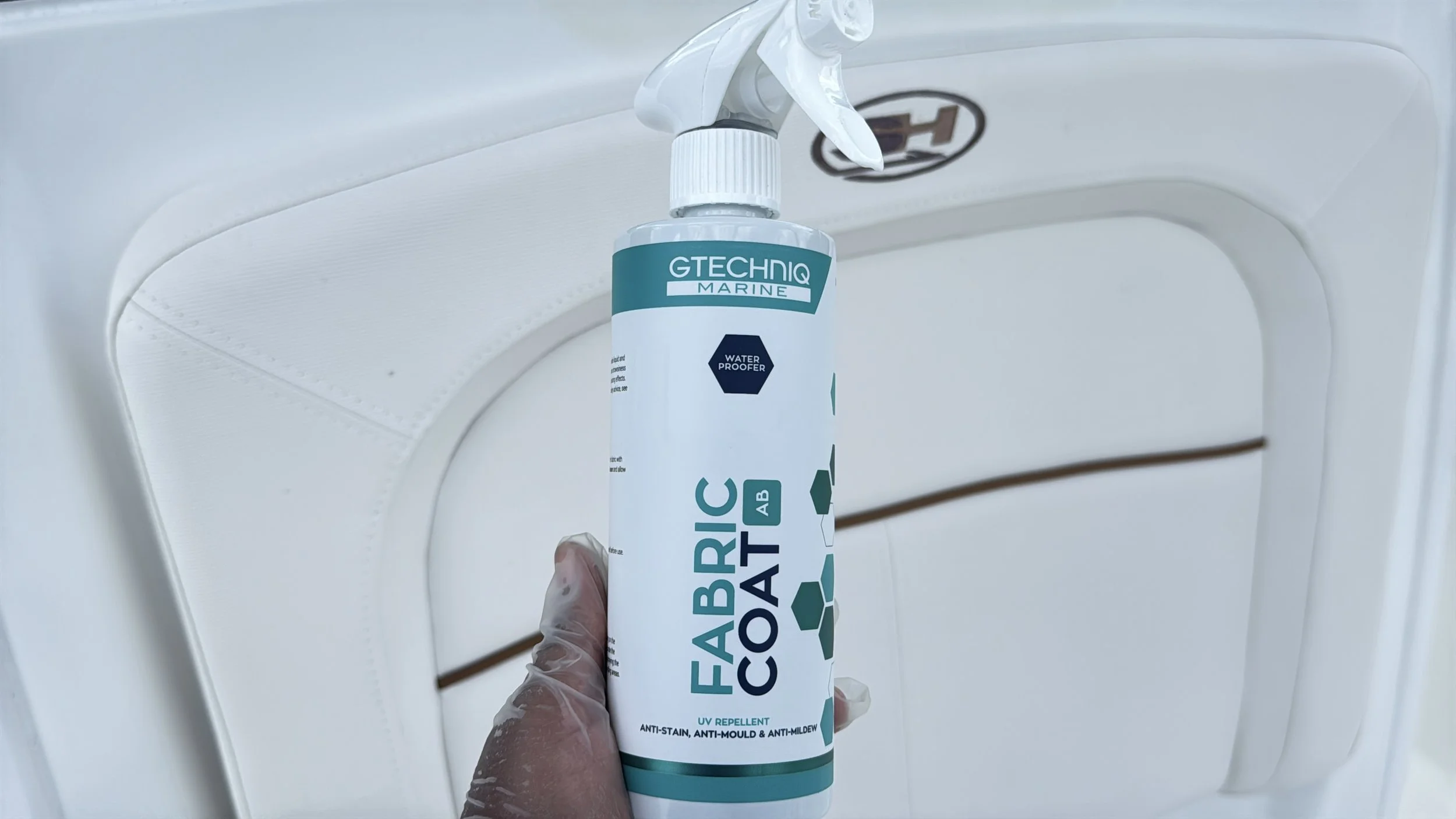 A person wearing a transparent glove holding a spray bottle of GTECHNIQ Marine Fabric Coat in front of a white washing machine.