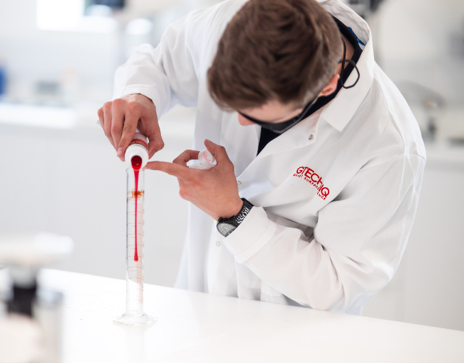 Scientist in a white lab coat pouring red liquid into a graduated cylinder in a laboratory.