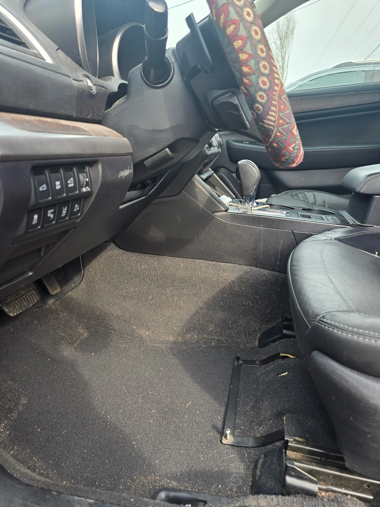 Inside view of a car's front interior showing the driver's side, gear shift, steering wheel with a patterned cover, and black seats with some wear, along with dust and debris on the carpet.