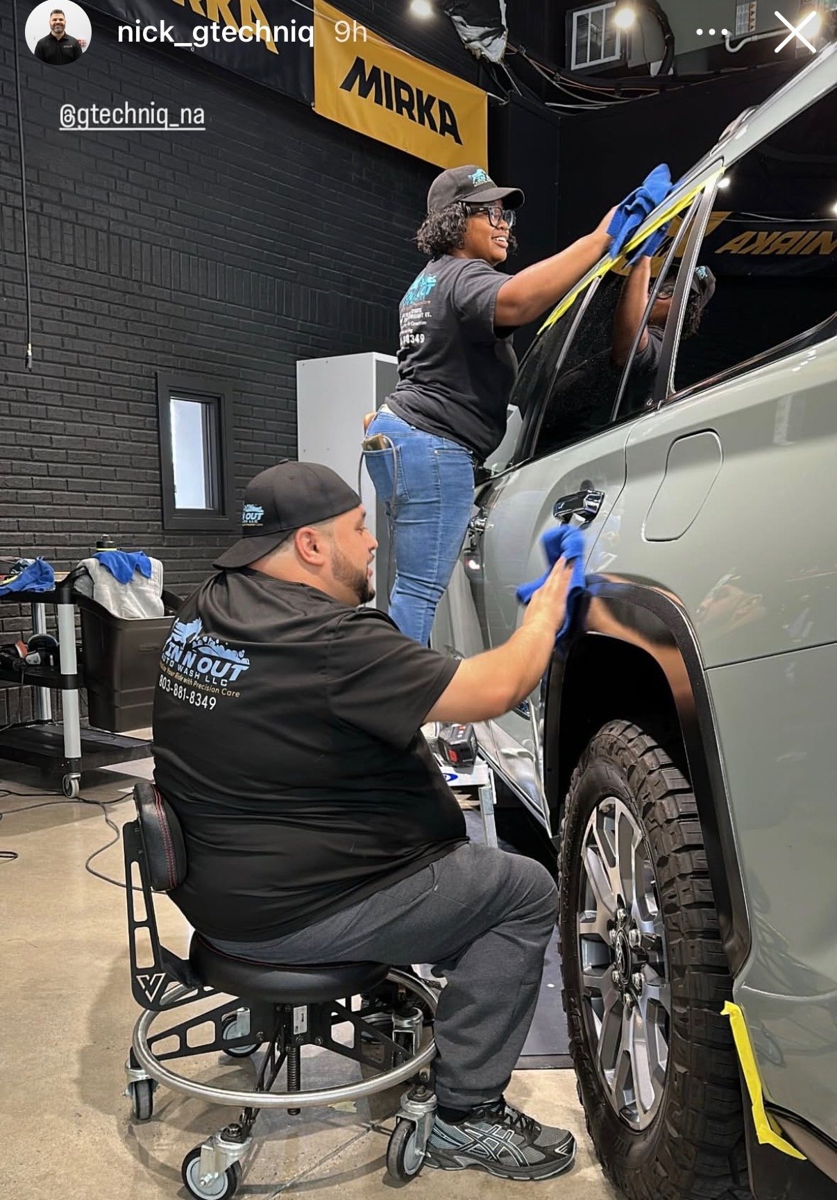 Two workers cleaning and detailing a silver SUV inside a workshop. The woman is standing on a step ladder wiping the top of the vehicle, while the man is sitting on a rolling stool polishing the side panel with a cloth.  "MIRKA" hanging from the ceil