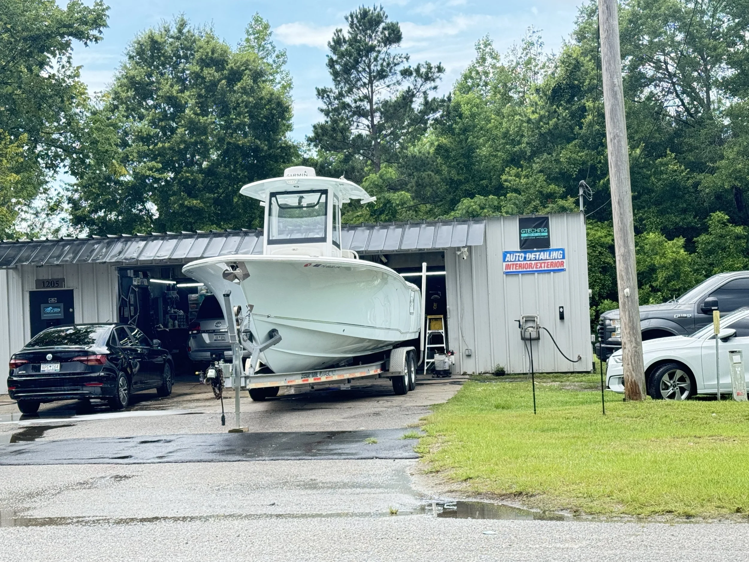 Auto detailing shop with a boat on a trailer, parked cars, and a sign that reads 'Auto Detailing Interior/Exterior' outside a gray building surrounded by green trees.