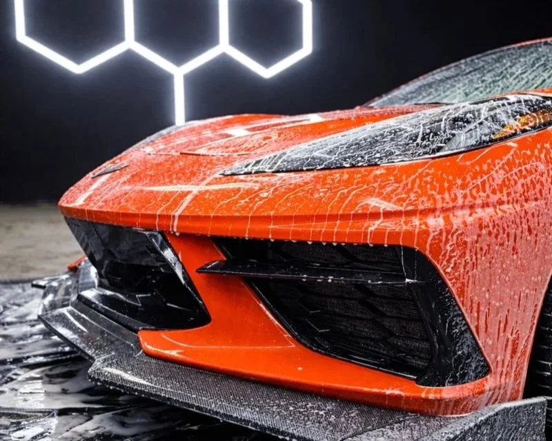 Close-up of a red sports car's front end, wet from water, with black accents and a carbon fiber front splitter, against a dark background with illuminated hexagonal light design.
