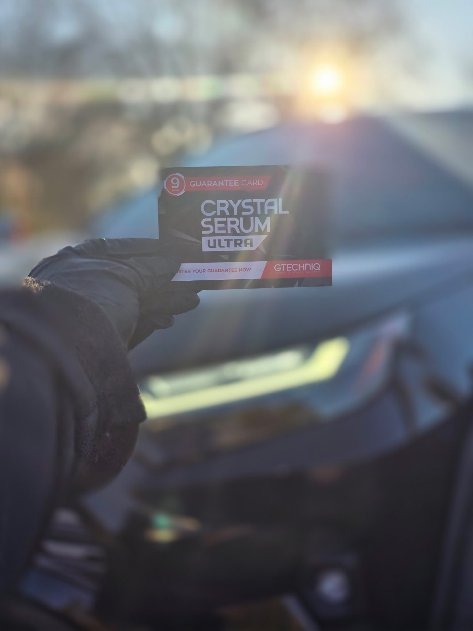 A hand in a black glove holds a small card labeled 'Crystal Serum Ultra' inside a vehicle, with sunlight shining through the windshield.