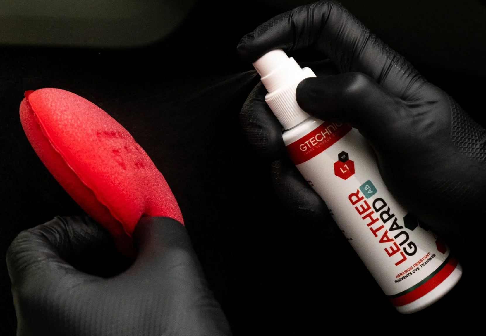 A person wearing black gloves uses a spray bottle of Leather Guard to treat a red car seat with a red microfiber application pad in their other hand.