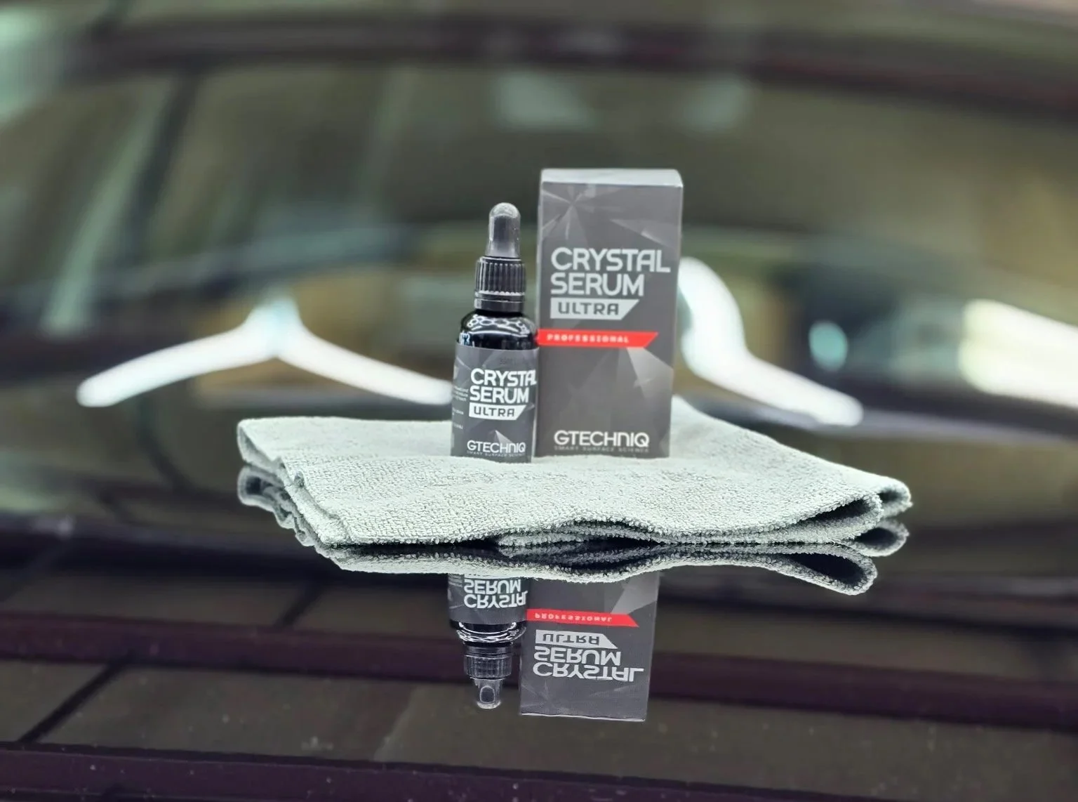 A bottle of Gtechniq Crystal Serum Ultra car coating with its box behind it, placed on a gray cloth on a car hood, reflecting on the shiny surface.