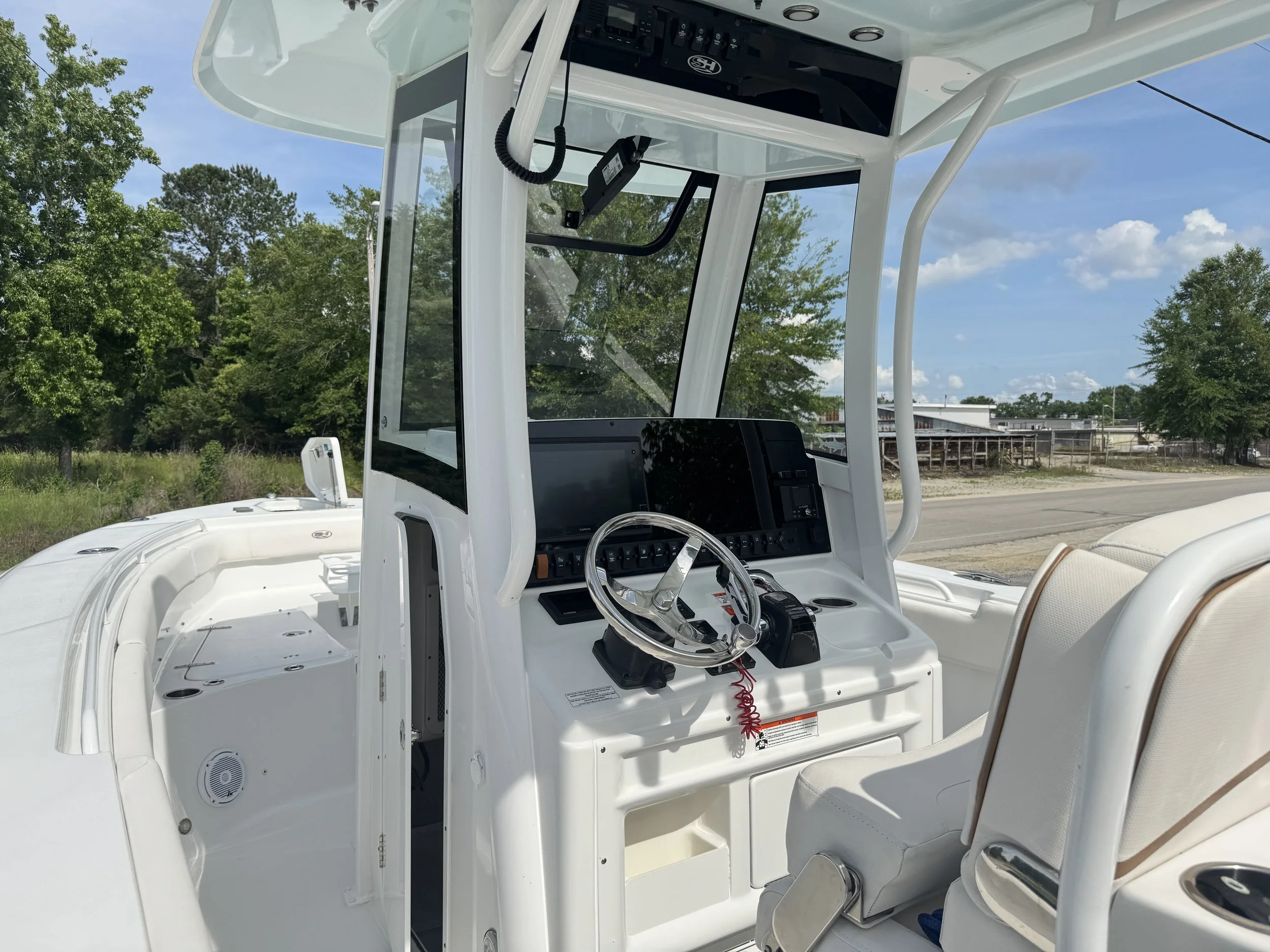Interior view of a white boat cockpit with steering wheel, control panel, and seating, docked outdoors with trees and blue sky in background.