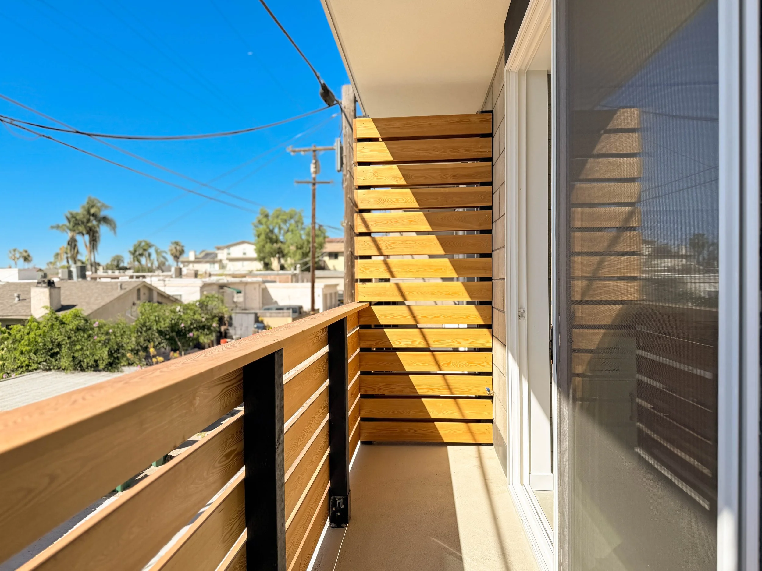 Small balcony with two wire chairs, a small round table with a potted plant, and a wooden privacy screen, overlooking a modern kitchen and living area through glass sliding doors.