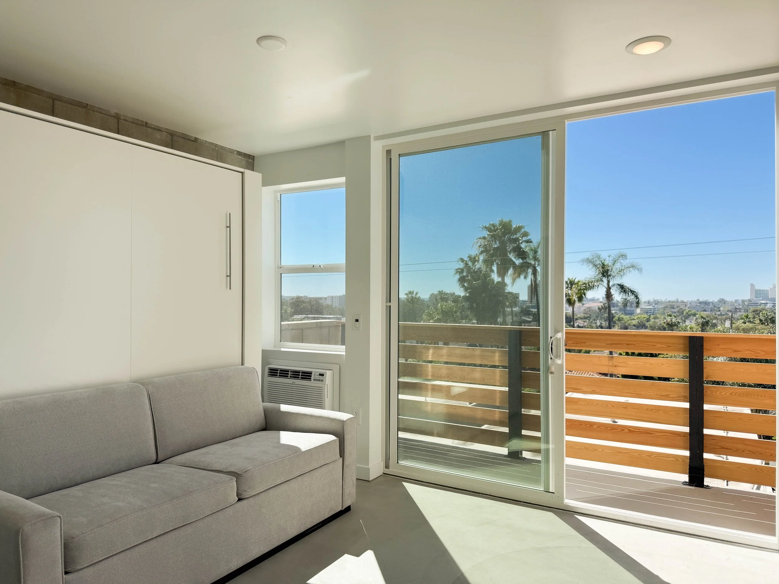Small apartment bedroom with a bed, small kitchen area, and balcony with wooden railing.