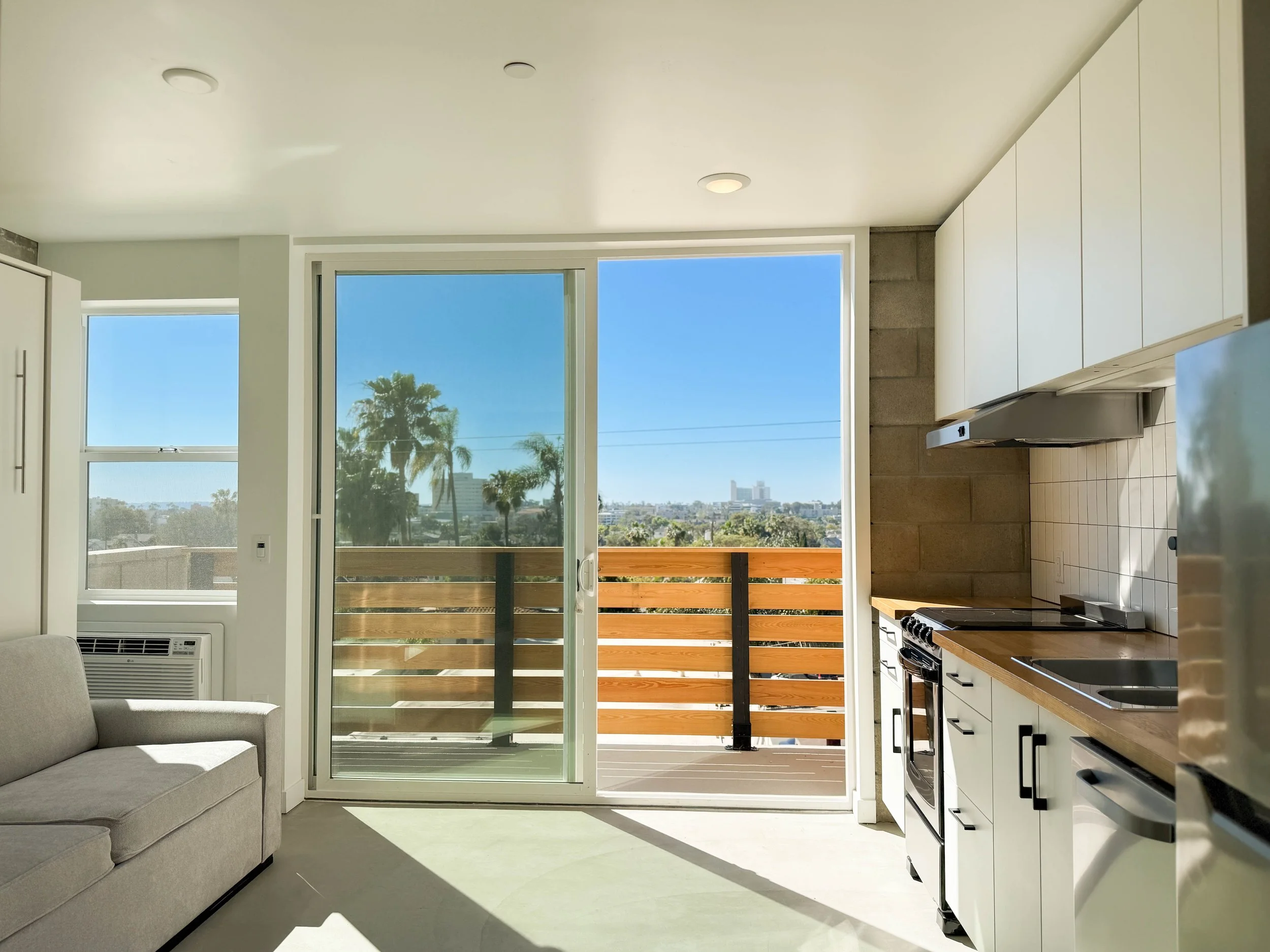 Living room with beige sofa, coffee table, sliding glass door leading to balcony with chairs and plant, modern decor, wood floors, and kitchen counter