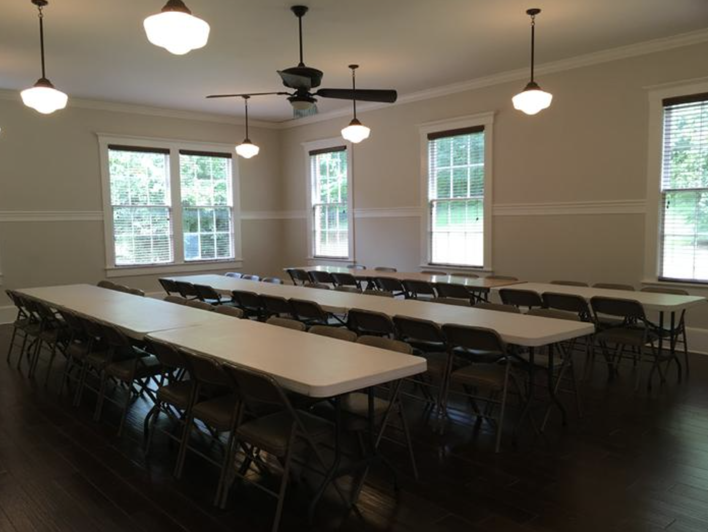 A spacious room with multiple long tables and chairs, illuminated by hanging ceiling lights and natural light from windows with blinds, has a ceiling fan and neutral-colored walls.