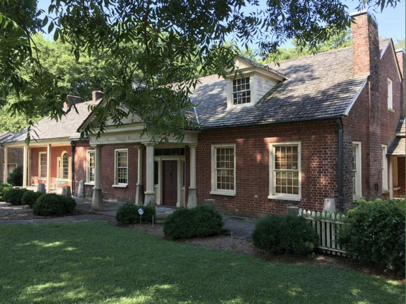 A two-story brick house with white trim, multiple windows, and a sloped shingled roof, surrounded by a well-kept lawn and shrubs, with large trees casting shadows.