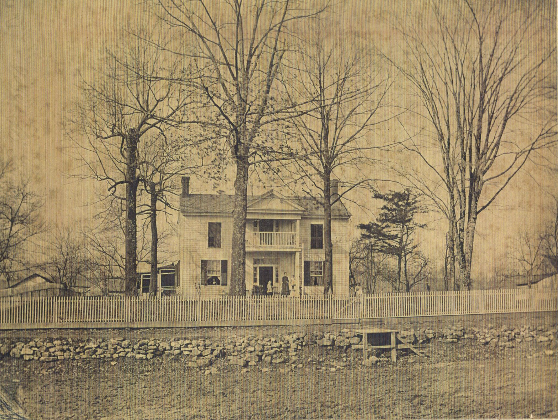 An old sepia-toned photograph of a large, two-story house with a porch, surrounded by tall leafless trees, and a white picket fence in the foreground.