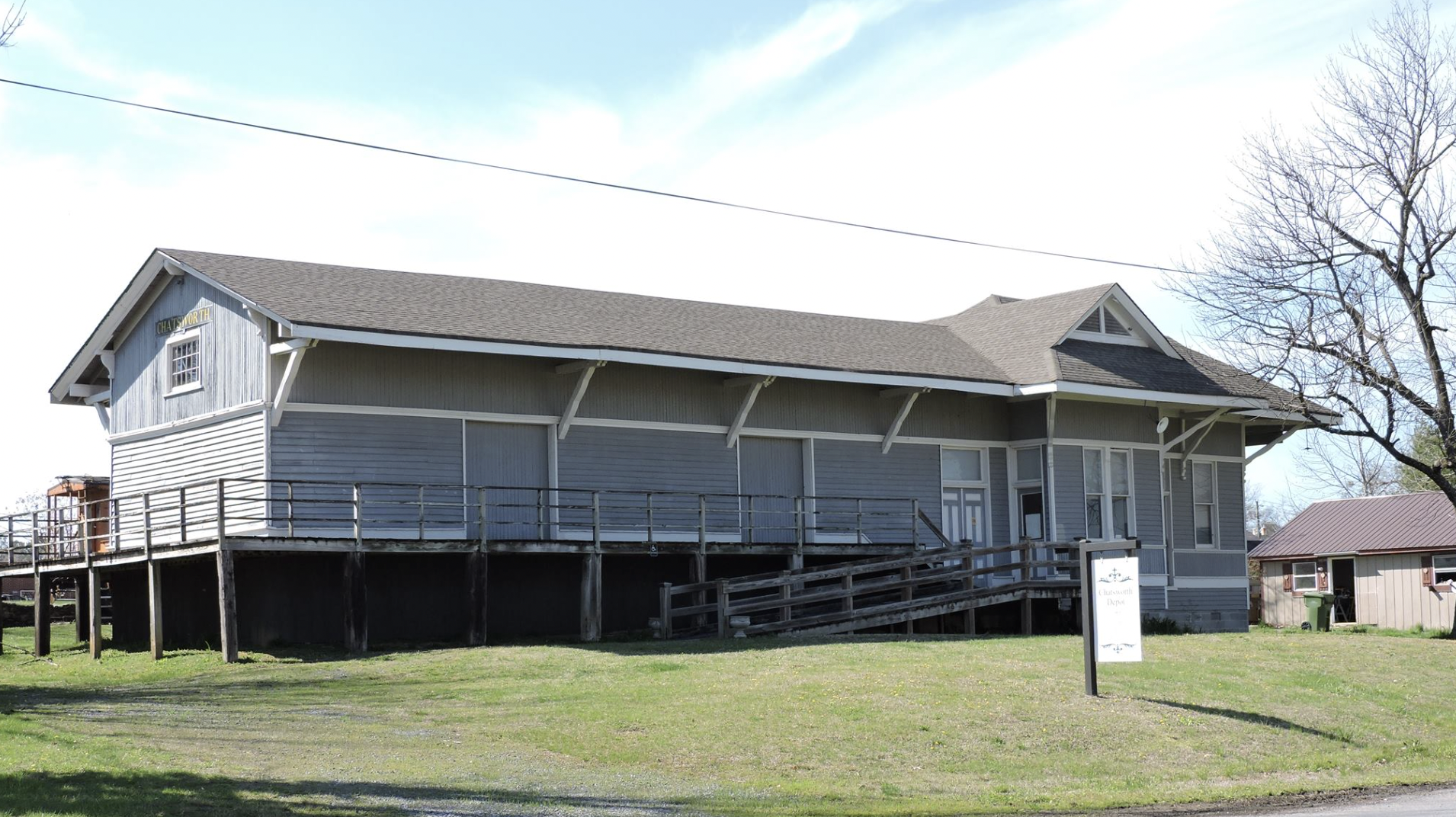 A light gray house with a porch, elevated on wooden stilts, with a ramp and steps leading to the entrance. The house has a gabled roof and multiple windows. There is a sign in front and a leafless tree on the right.