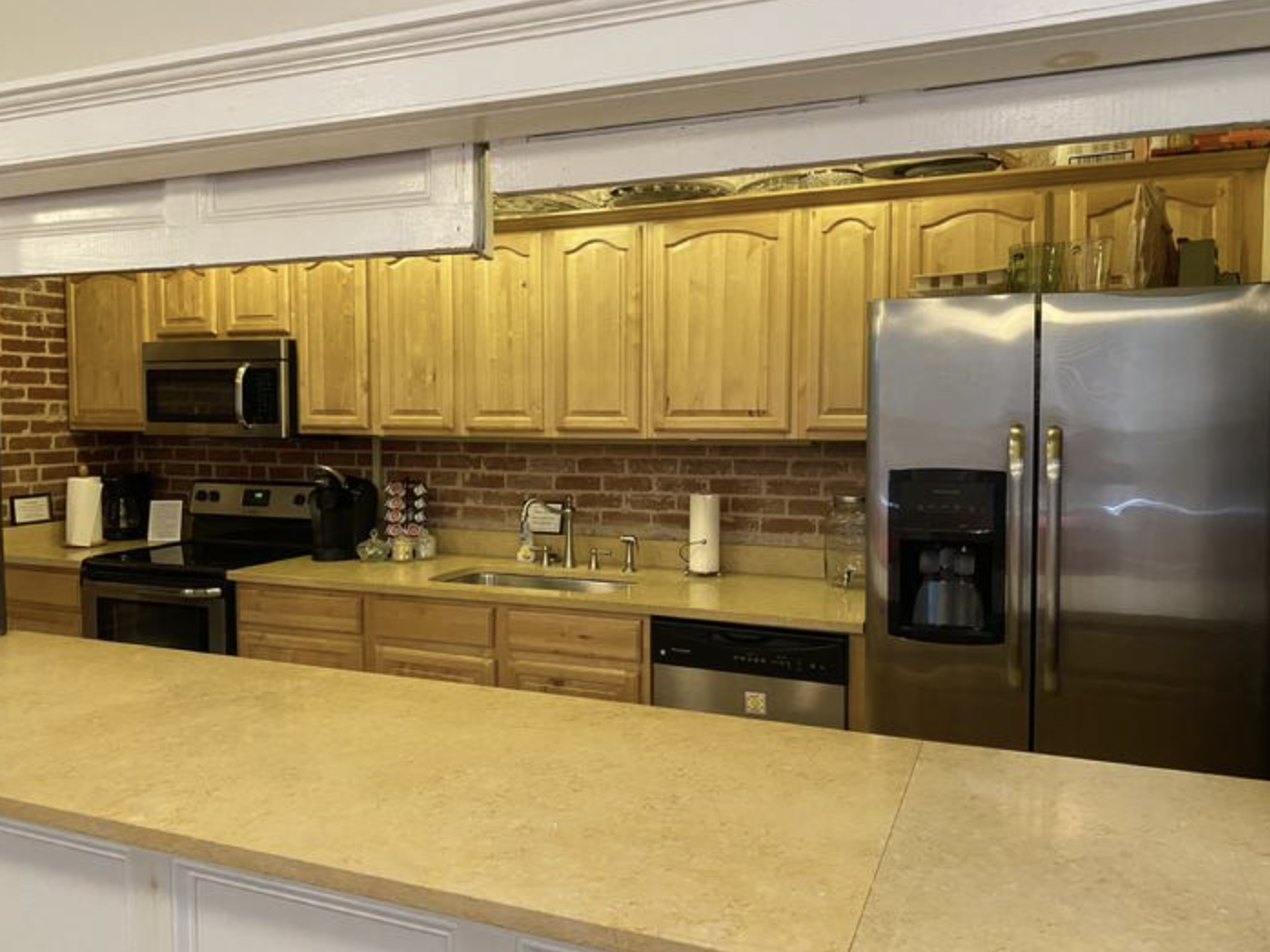 A kitchen with wooden cabinets, stainless steel refrigerator, microwave, and black appliances, brick wall backsplash, and beige countertop, viewed through a white opening.
