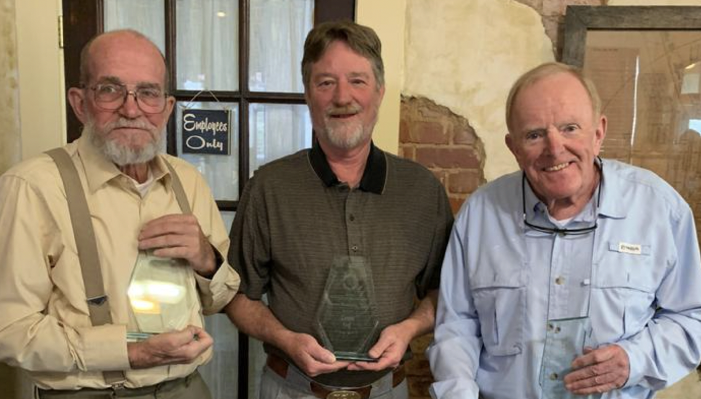Three elderly men smiling, standing together inside a rustic restaurant or café. The man on the left holds an award or plaque, the man in the middle holds a glass award, and the man on the right holds a small object. Behind them is a sign that reads 'Employees Only' and a brick and wood interior.