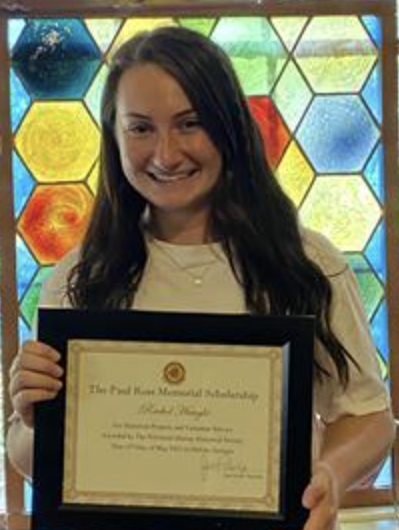 Young woman smiling and holding a framed certificate in front of a colorful stained glass window.
