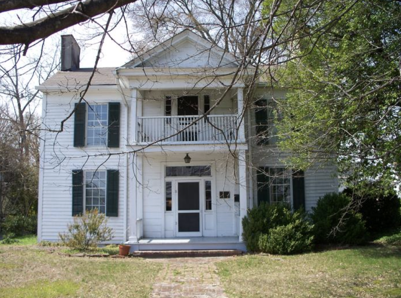 Front view of a two-story white house with green shutters, a porch, and a balcony, surrounded by trees and shrubs.