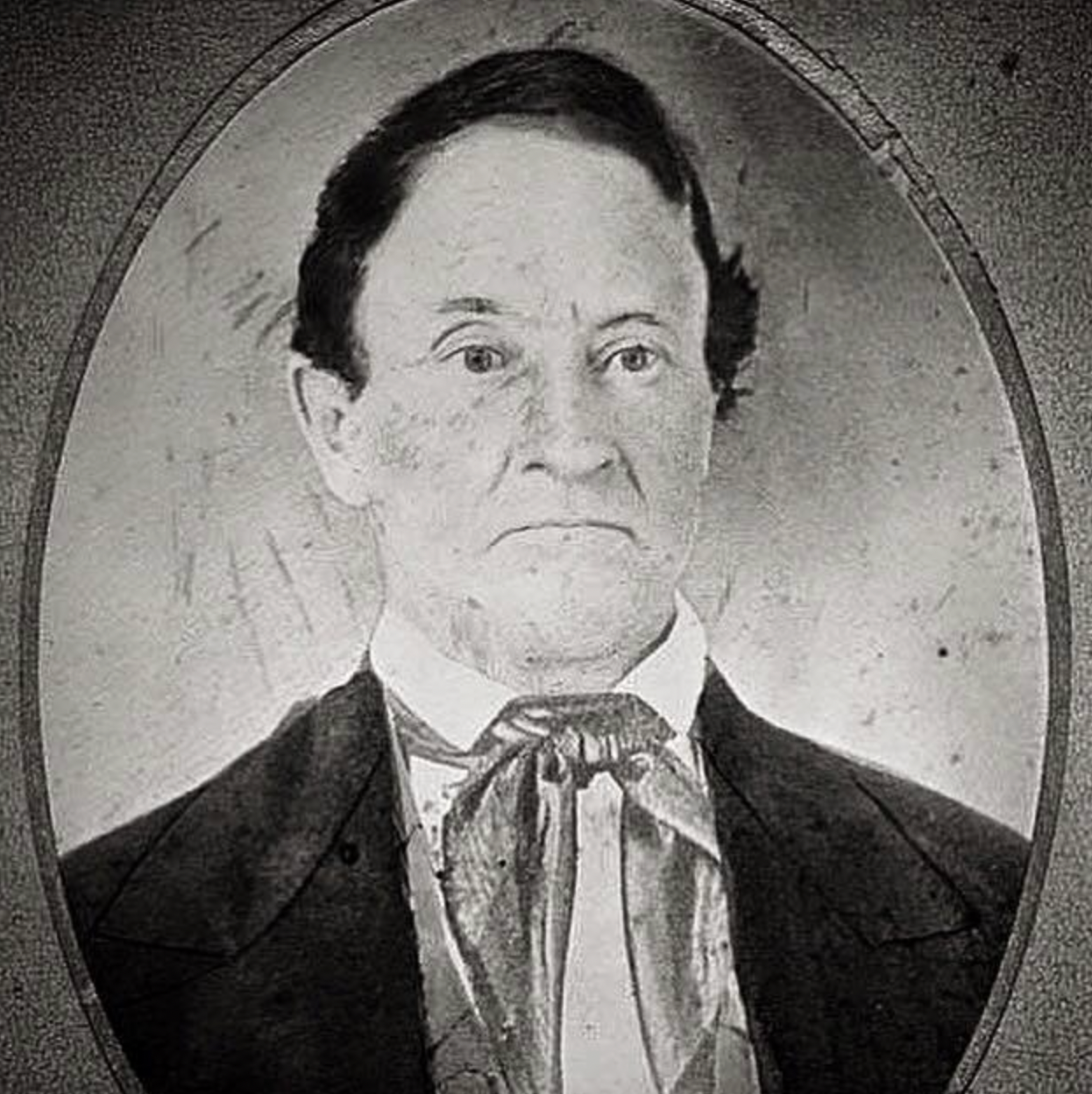 Black and white photograph of a serious-looking man with dark hair, dressed in historical attire, including a cravat or necktie, within an oval frame.