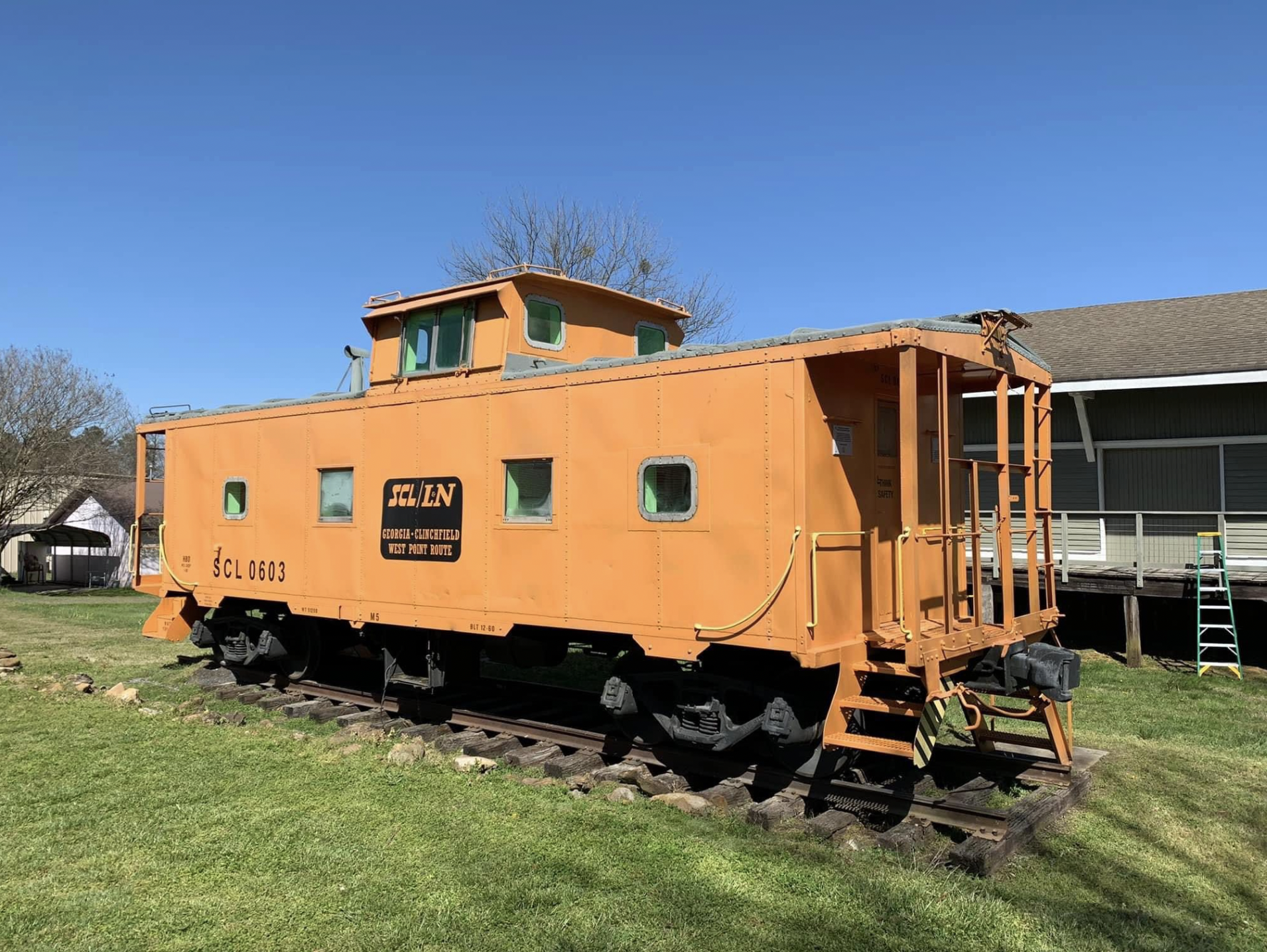 Orange vintage train caboose on a grassy area with a blue sky, trees, and a building in the background.