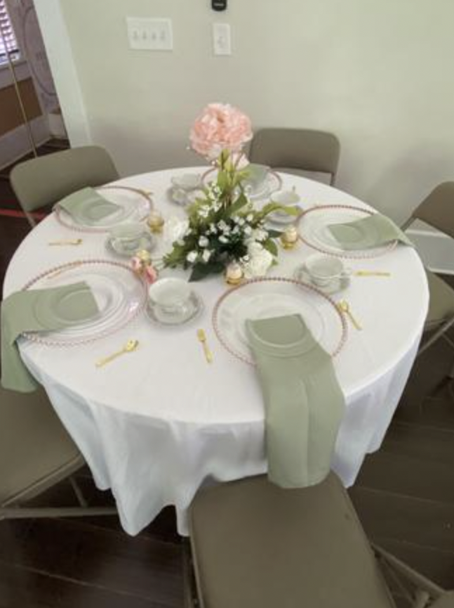 Round table set for six with a white tablecloth, green napkins, gold cutlery, and a pink floral centerpiece with white accents.