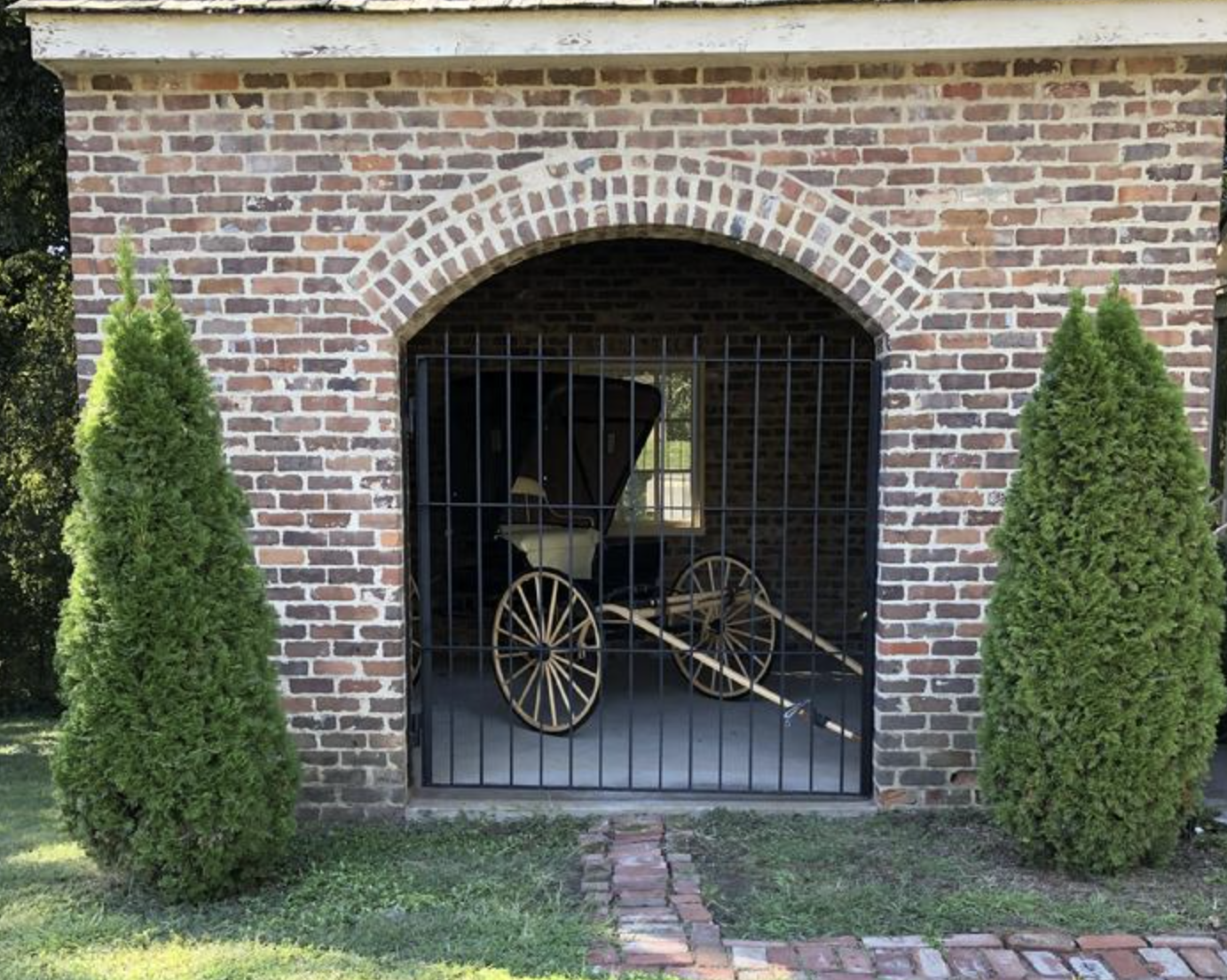 A vintage baby carriage is stored behind black metal bars within a brick archway, flanked by two small green shrubs outside.