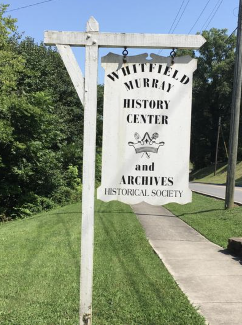Sign for Whitfield Murray History Center and Archives, located along a sidewalk on a grassy area with trees in the background.