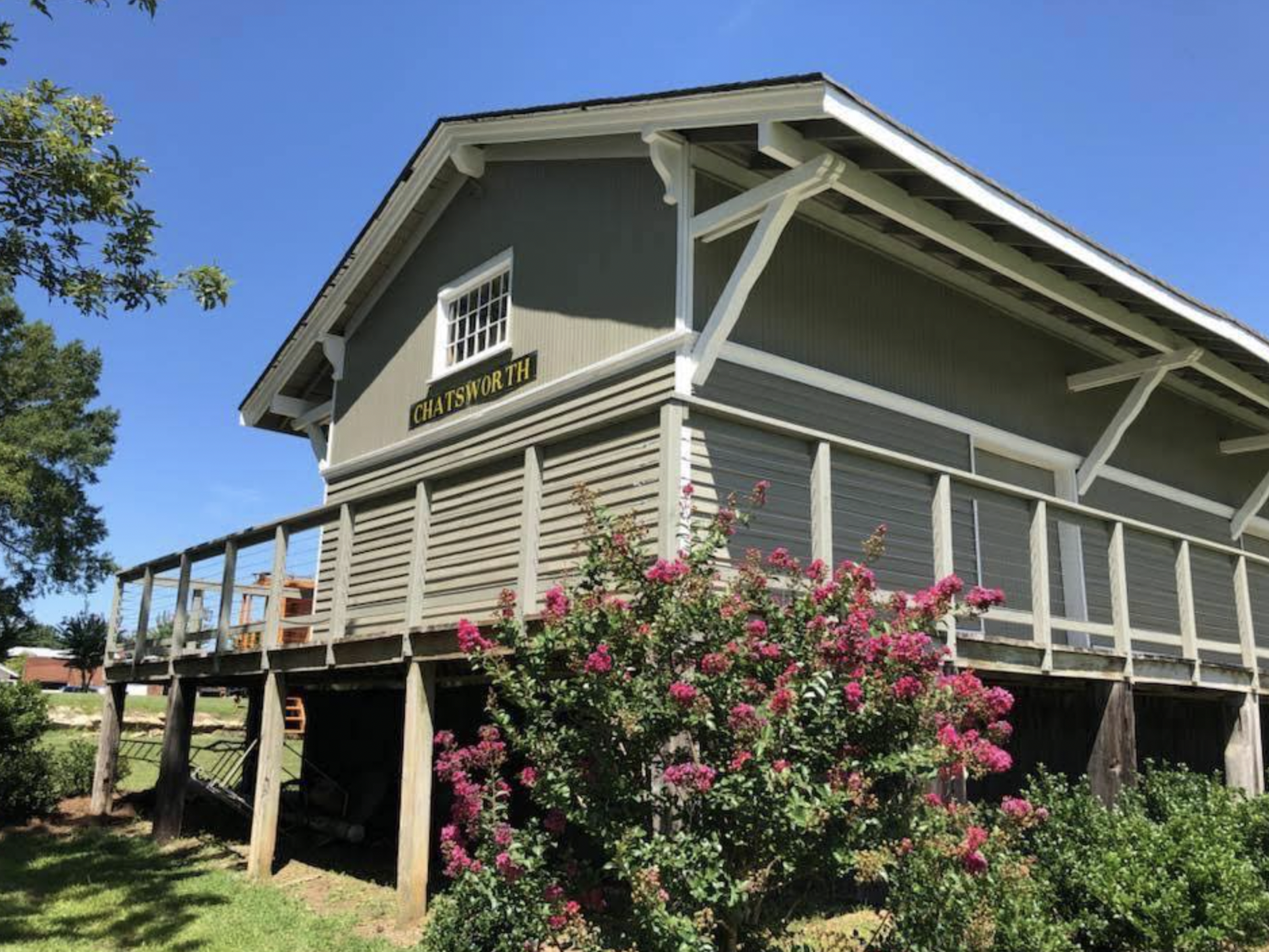 A raised train station house with a sign labeled 'Chatsworth', surrounded by green grass, pink flowering shrubbery, and trees, under a bright blue sky.