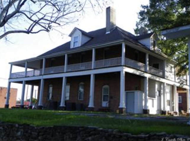 A large, two-story house with a wraparound porch, brick and white siding, and a steep roof with dormer windows. There is a grassy lawn in front and some trees around the house.
