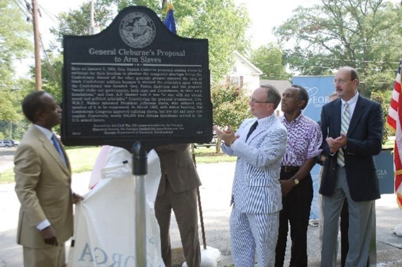 A group of men in formal and semi-formal attire at a historical marker unveiling ceremony outdoors during daytime, with trees and American flags in the background.
