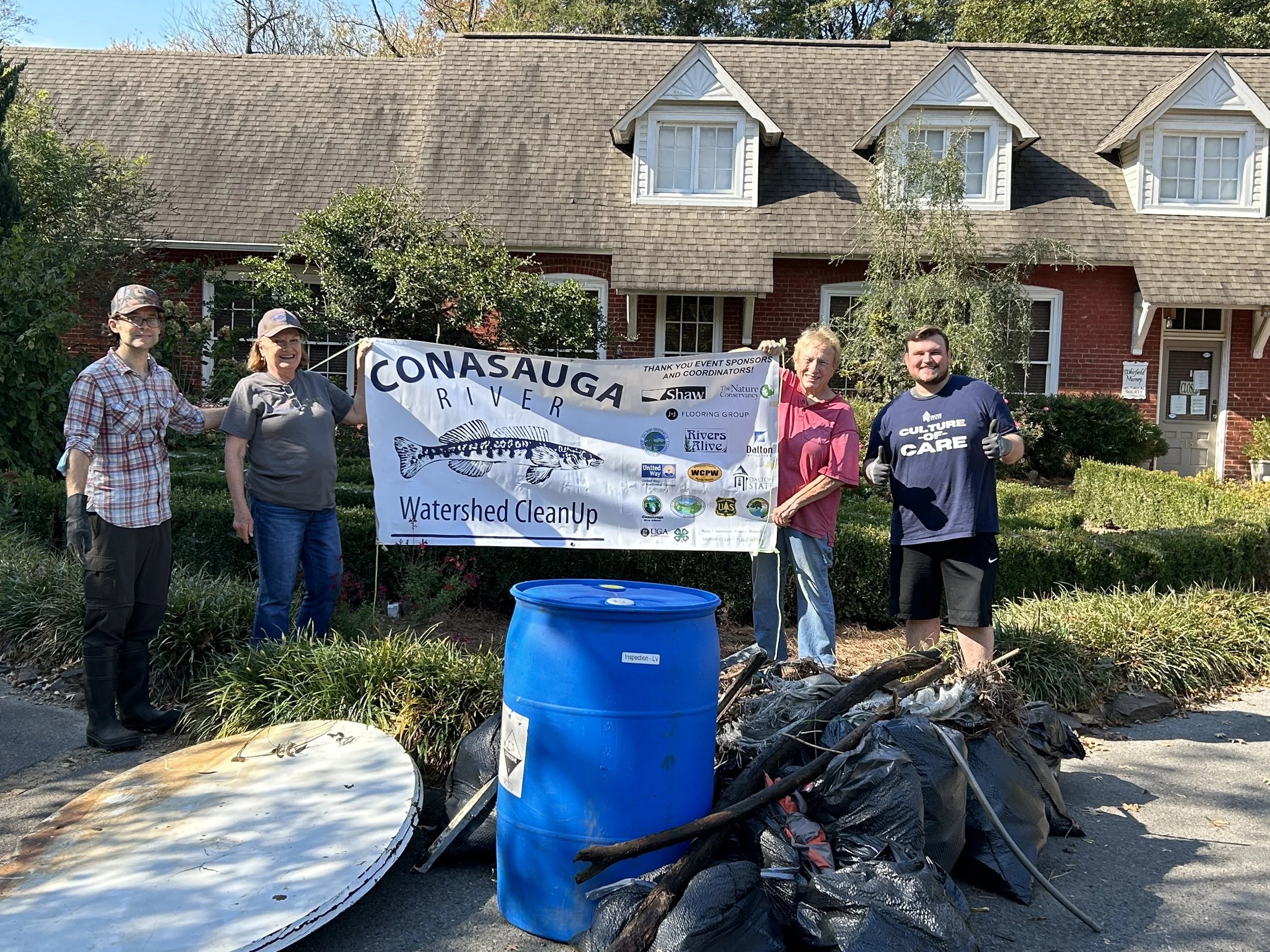 Group of five people standing outdoors, holding a banner that reads 'CONASAUQA RIVER Watershed CleanUp,' with trash and debris in front of them, in a residential neighborhood with a brick house in the background.