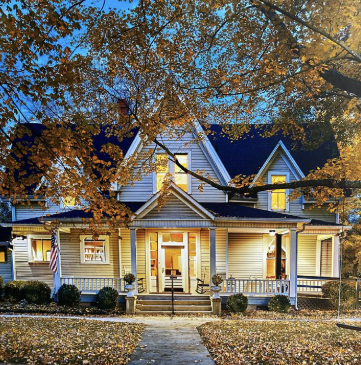 A large, white, two-story house with a front porch and multiple gables, surrounded by trees with yellow leaves, during fall.