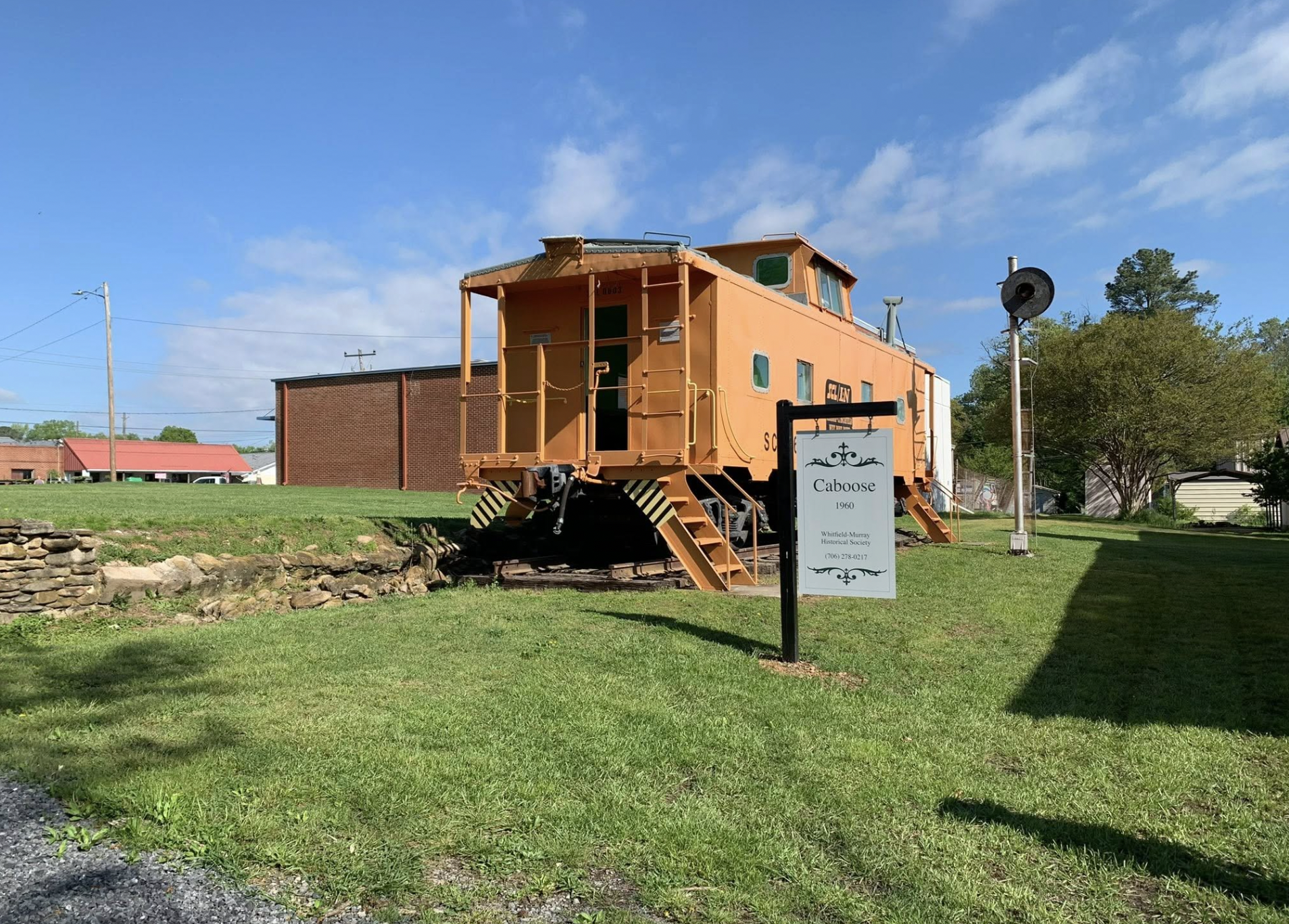 Vintage orange caboose train car on display outdoors, with a sign reading "Caboose 1960" from the Whitefield-Murray Historical Society, green grass, a stone retaining wall, clear blue sky with a few clouds.