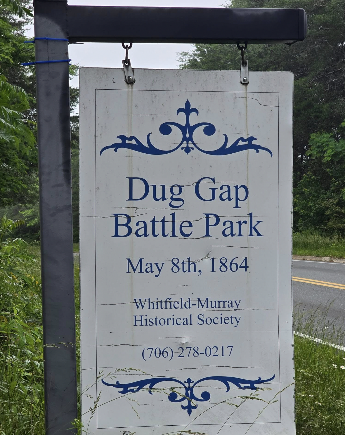 Sign for Dug Gap Battle Park with date May 8th, 1864, provided by Whitfield-Murray Historical Society, with a phone number, near a road and green trees.