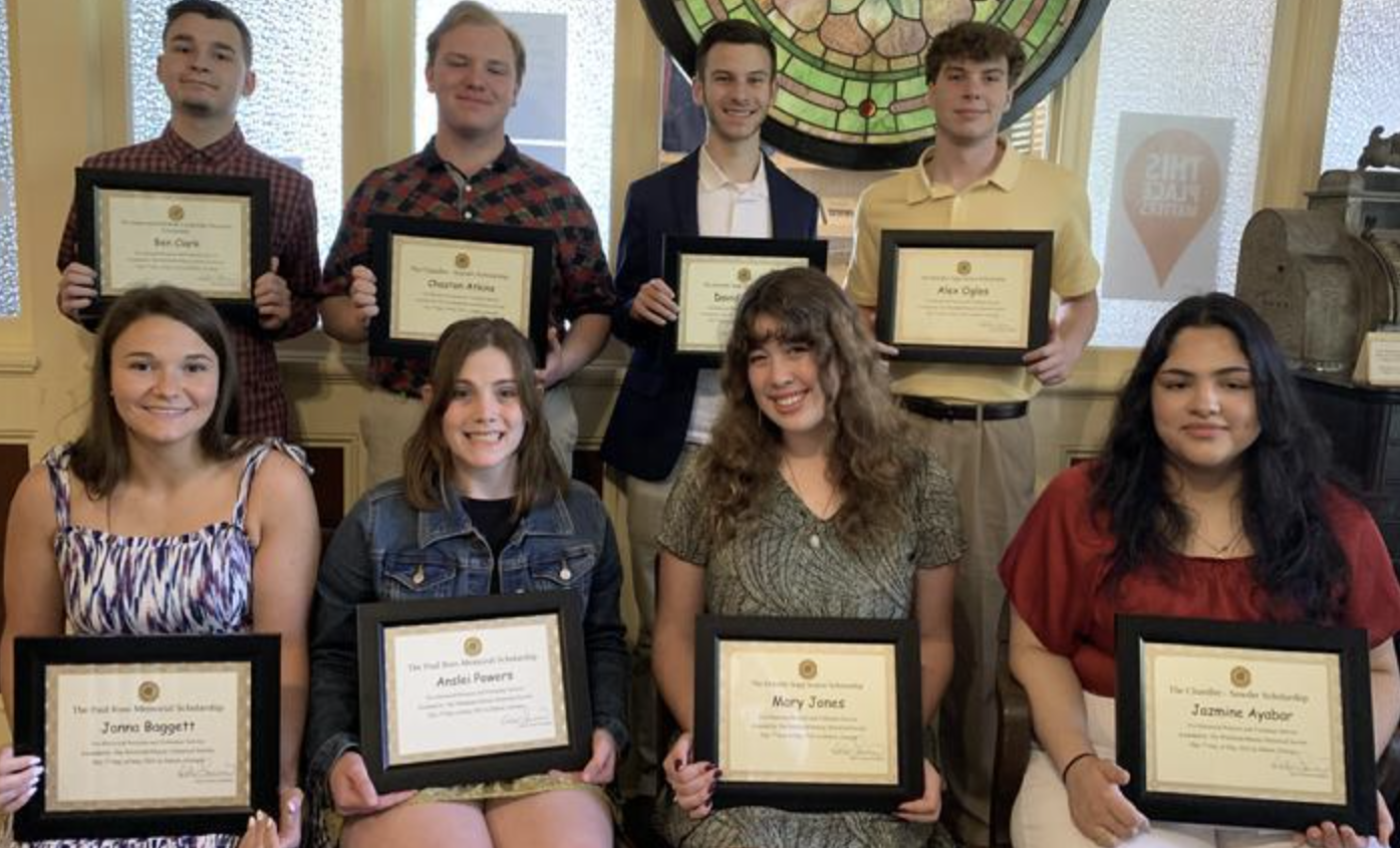 Group of nine teenagers, five standing in the back and four sitting in the front, holding certificates of achievement or awards in a room with light-colored walls and windows, celebrating their accomplishments.