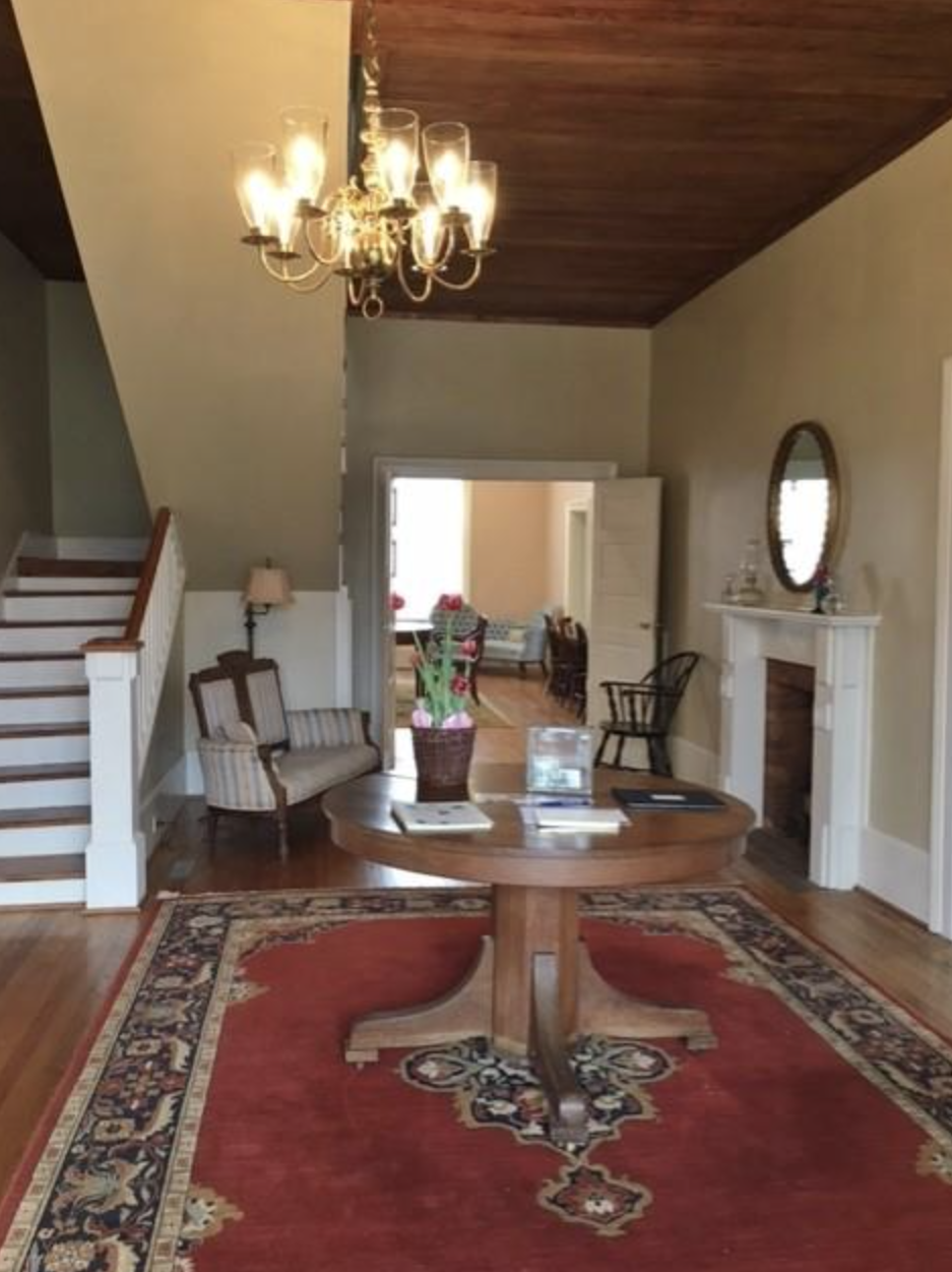 Living room with a round wooden table on a red patterned rug, a chandelier hanging from the ceiling, a fireplace with a mirror above it, a small bench, and a doorway leading to another room with chairs and a window.