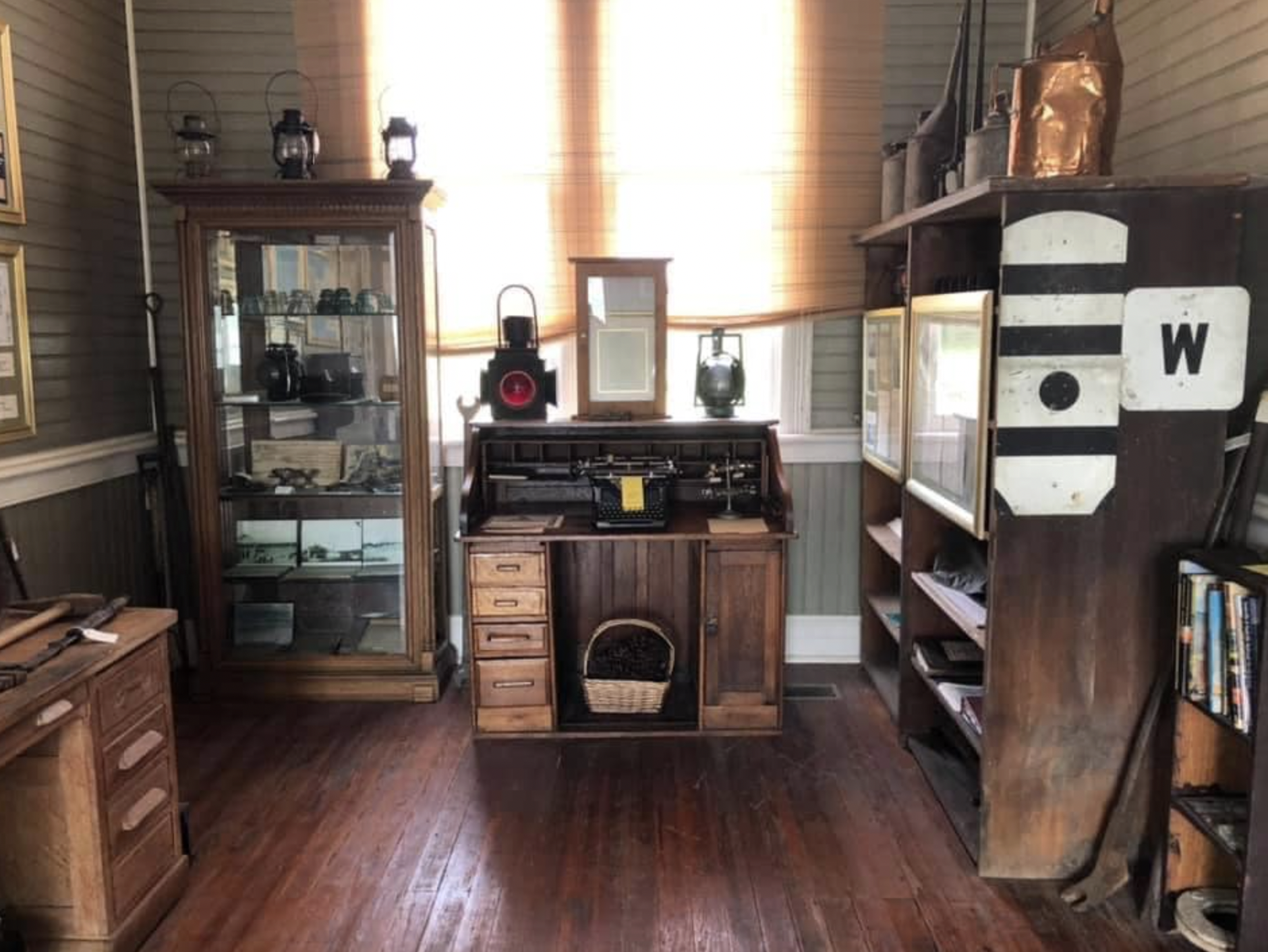 Room with wooden furniture and display cases, containing vintage lanterns, old books, and collectibles