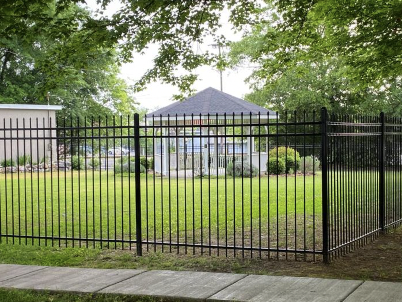 A black metal fence surrounds a well-maintained grassy area with a small gazebo in the background, trees overhead, and some buildings and parked cars partially visible beyond the fence.