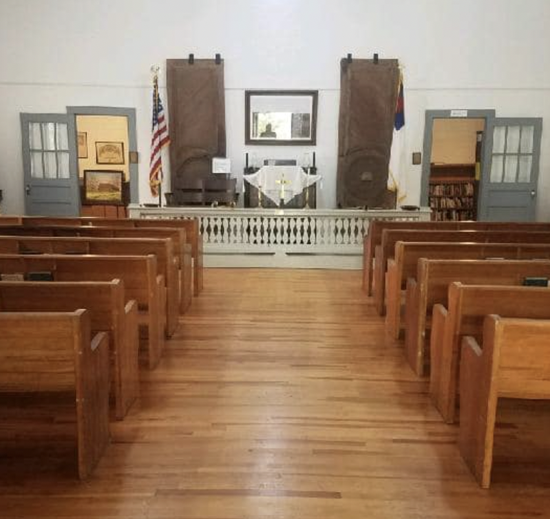 Inside a historic church with wooden pews facing an altar, flags, and two large wooden chairs.