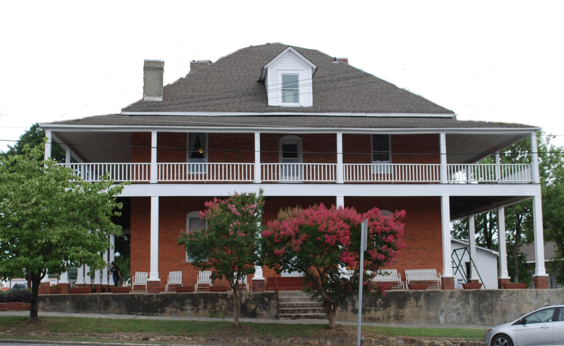 A large red brick house with a spacious wrap-around porch, white railings, and multiple windows, surrounded by green trees and colorful flowering bushes.