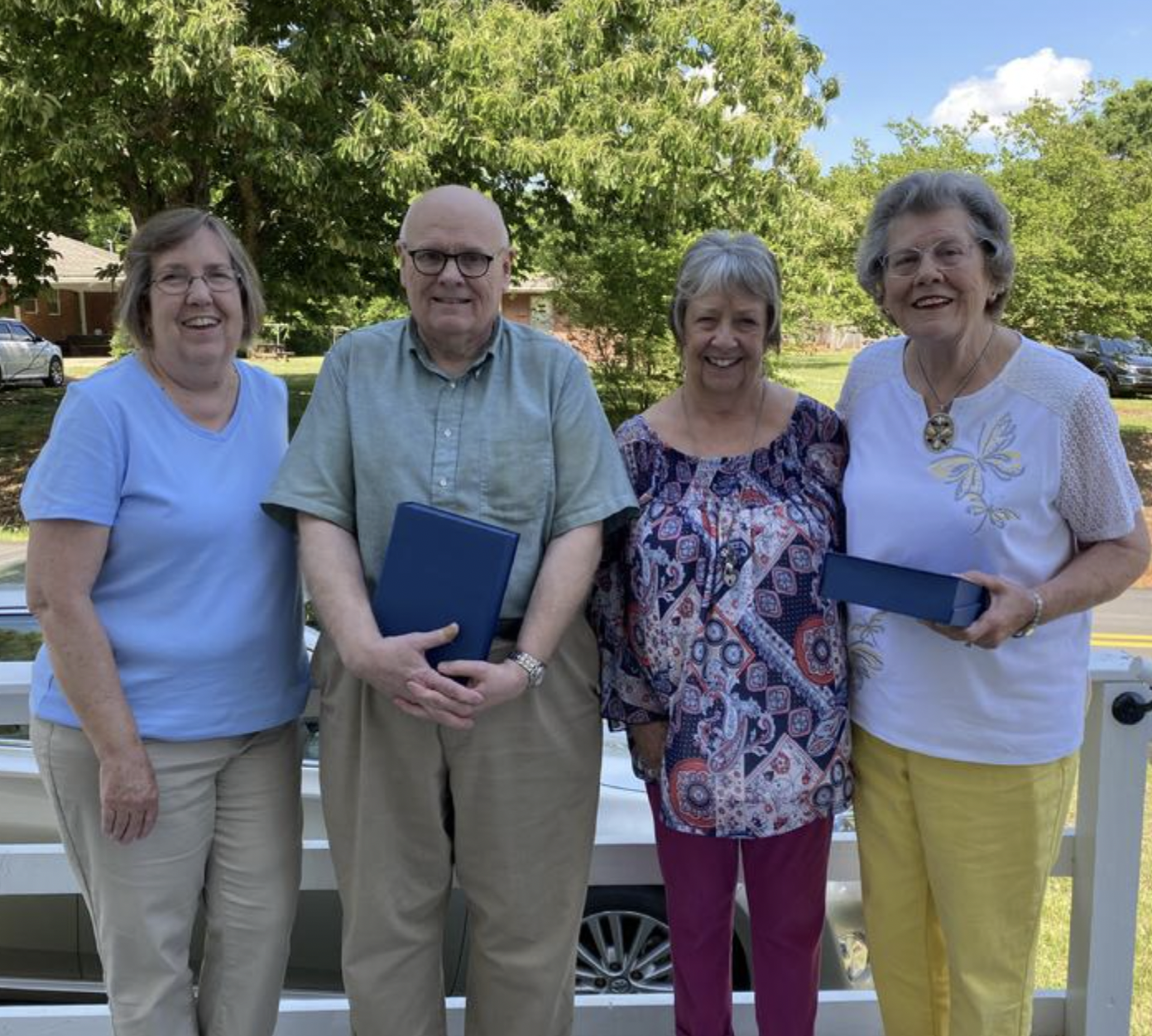 Four older adults standing outdoors on a sunny day in front of a white fence, holding blue boxes. Two women are wearing glasses, and all are smiling. There are trees and houses in the background.