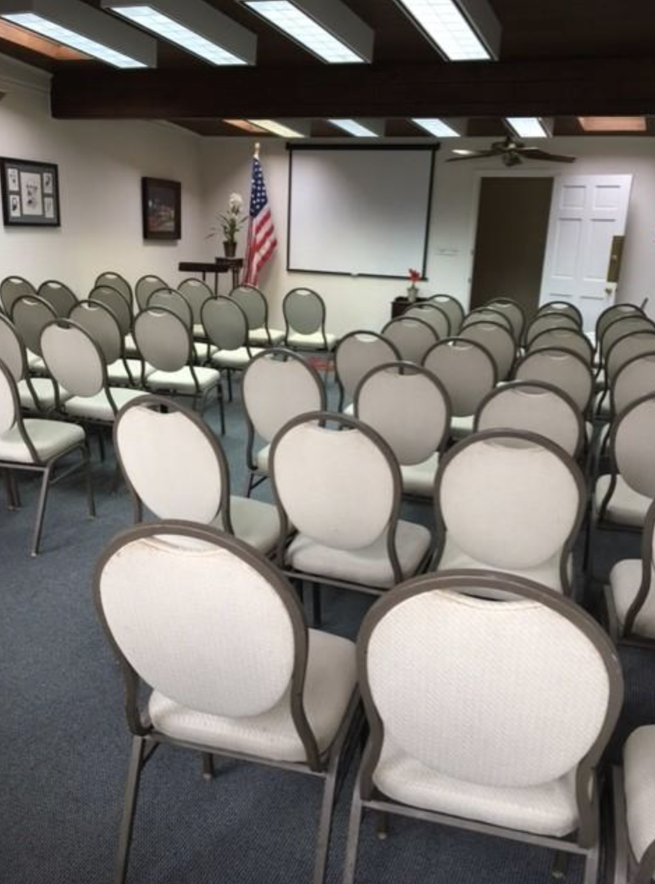 An empty conference or meeting room with multiple rows of white chairs with black frames, a projector screen, a ceiling fan, and an American flag in the corner.