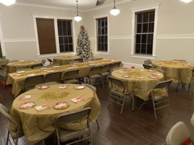 Room decorated for a holiday party with a Christmas tree in the corner, round tables with yellow tablecloths, red and white plates, and napkins, and chairs around the tables.