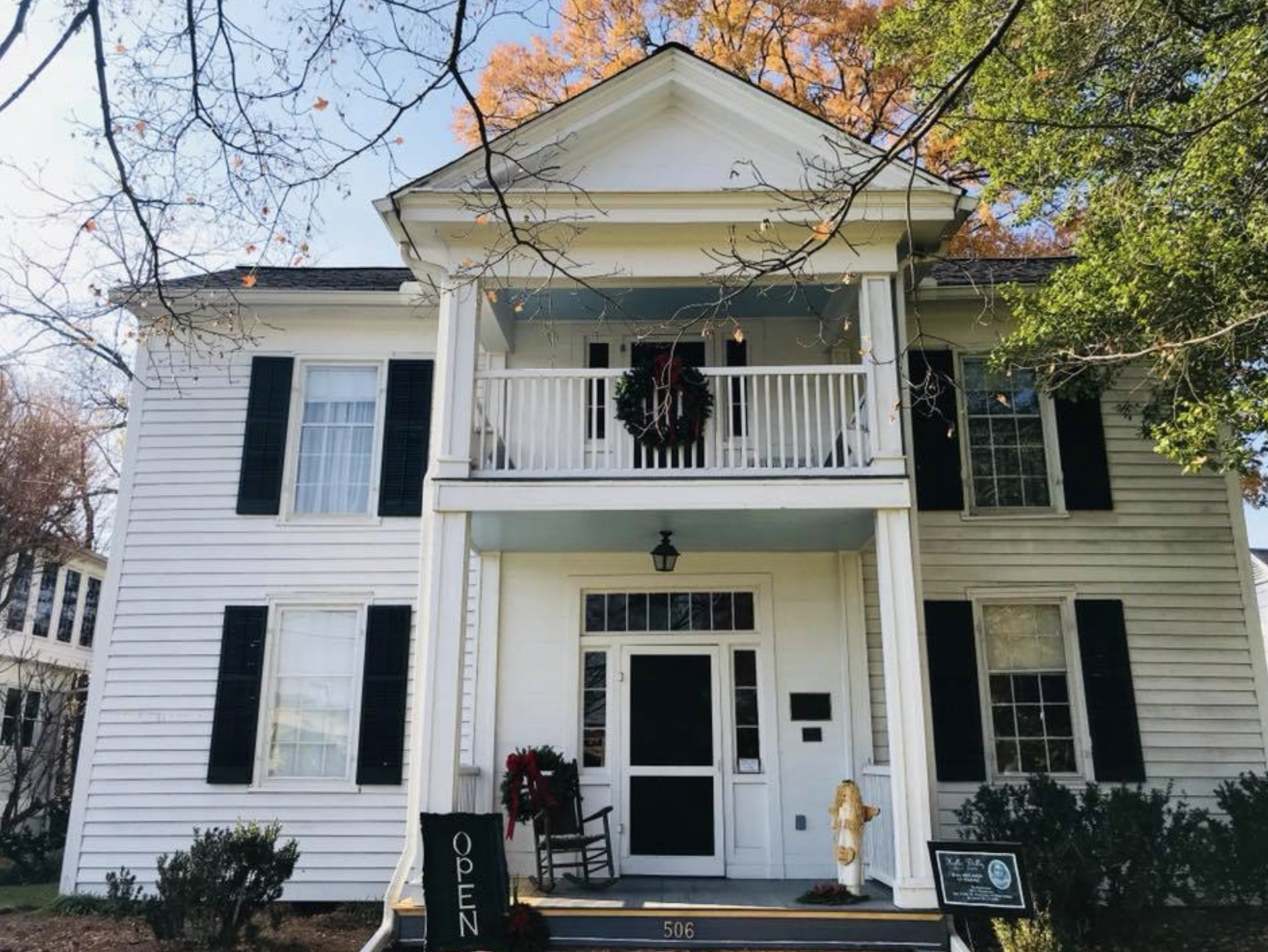 White two-story house with black shutters, decorated with a wreath on the porch, and signs indicating it is open for viewing, set against a backdrop of trees with autumn leaves.